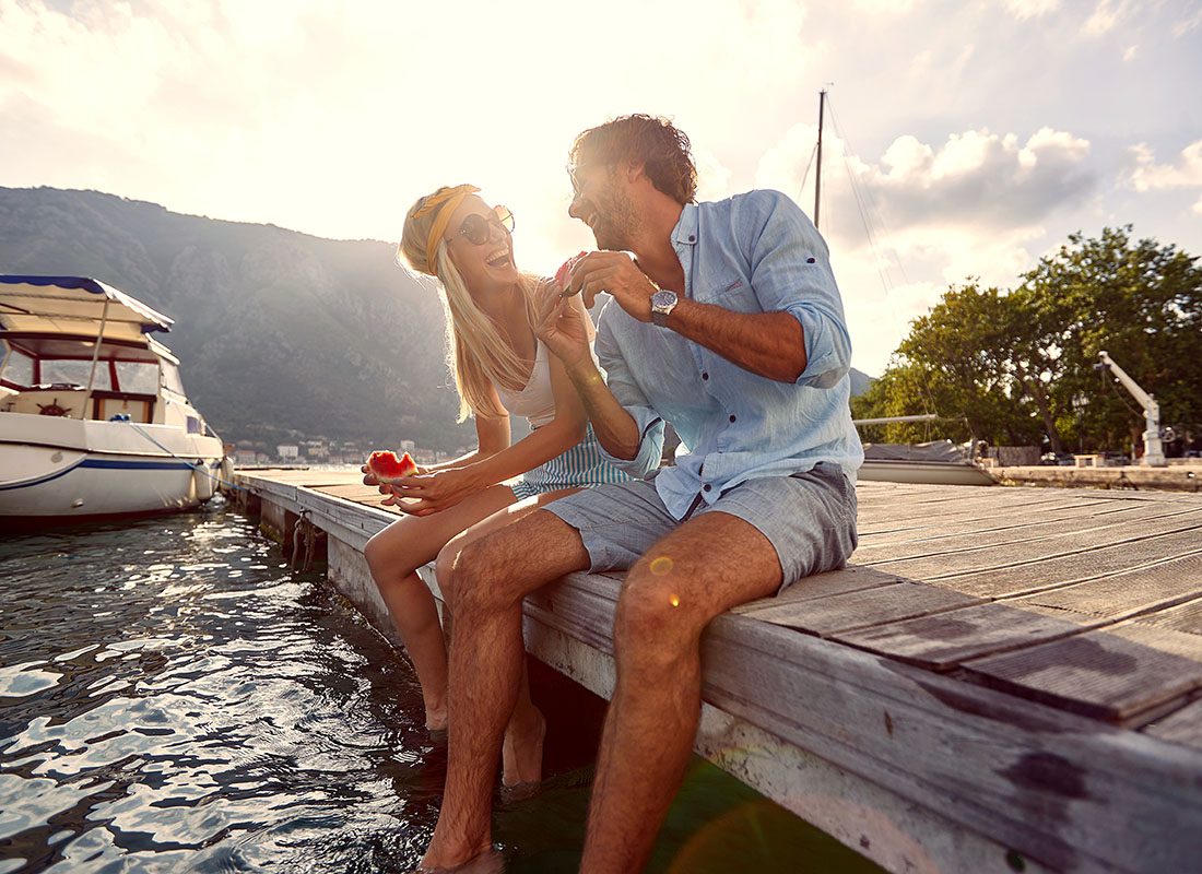 Read Our Reviews - Portrait of a Cheerful Young Couple Having Fun Sitting on a Wooden Dock at the Lake Near a Boat While Eating Watermelon During a Summer Trip